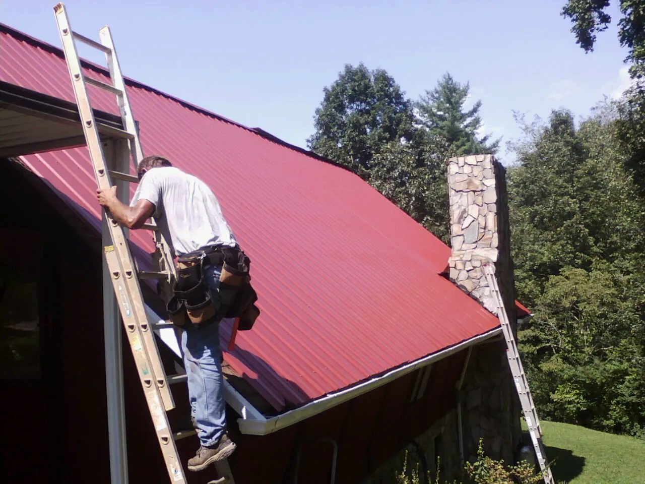 Roofer on a ladder beside a red metal roof and stone chimney — Soffit & Fascia Repair by Roofing Brothers in Kooskia